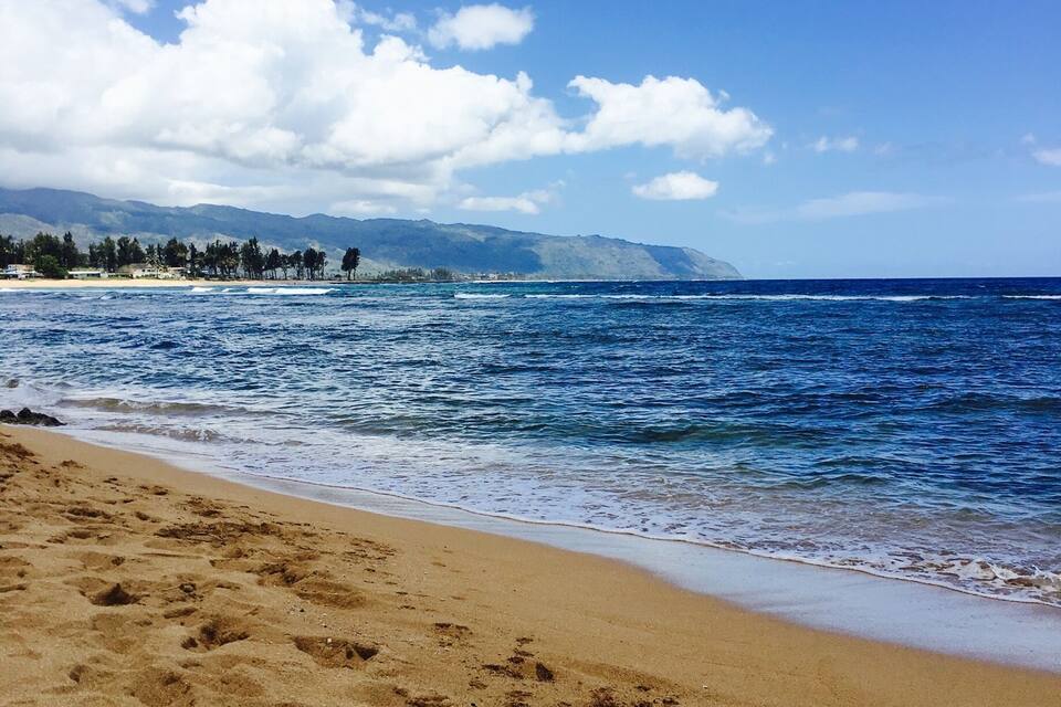 Uma praia com águas azul-claras, areal arenoso e montanhas ao longe.