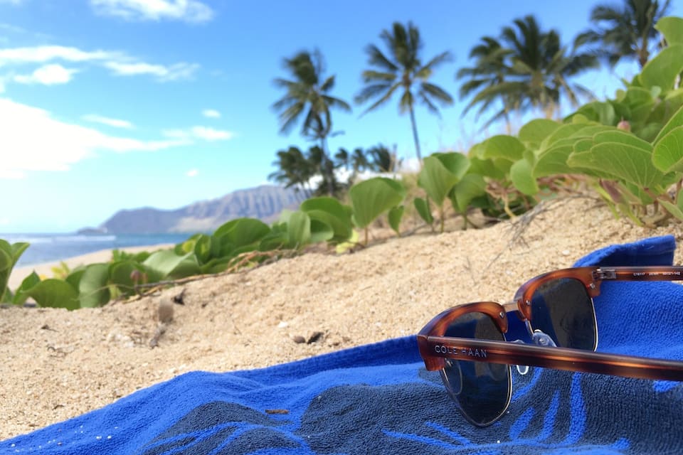 Sunglasses on a blue towel on a sandy beach with palm trees and a mountain in the background.