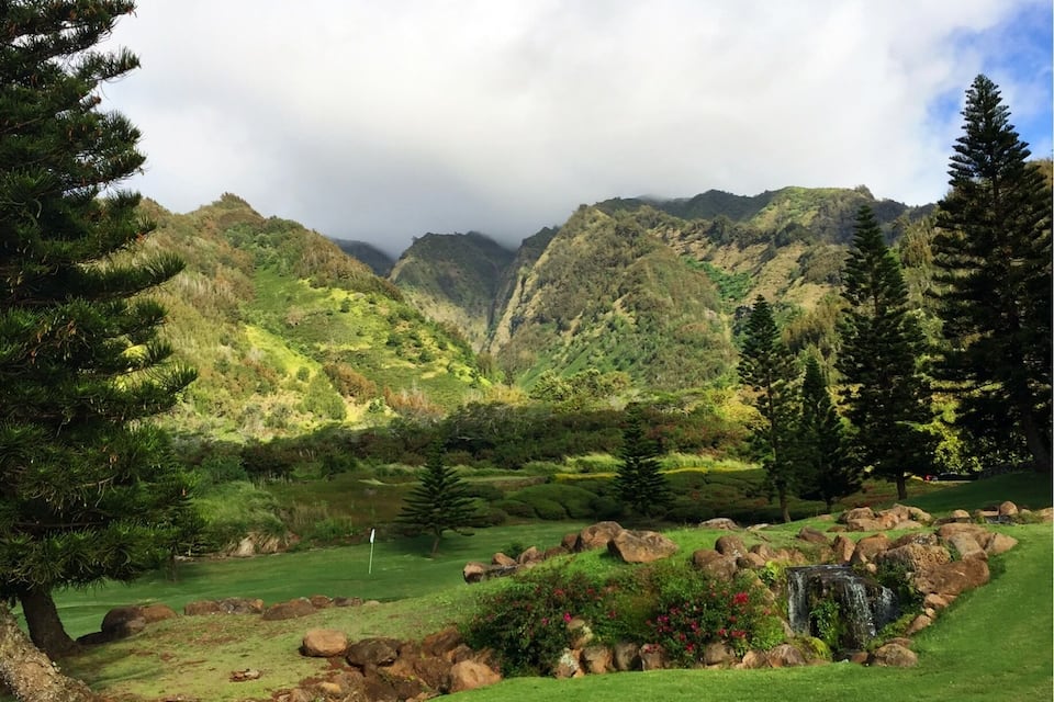 Ein üppiger Golfplatz mit einem Wasserfall, umgeben von Bergen und Kiefern.