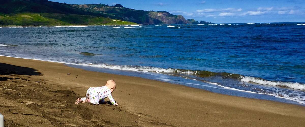 A baby on a sandy beach with waves crashing onto the shore and a hilly landscape in the background.