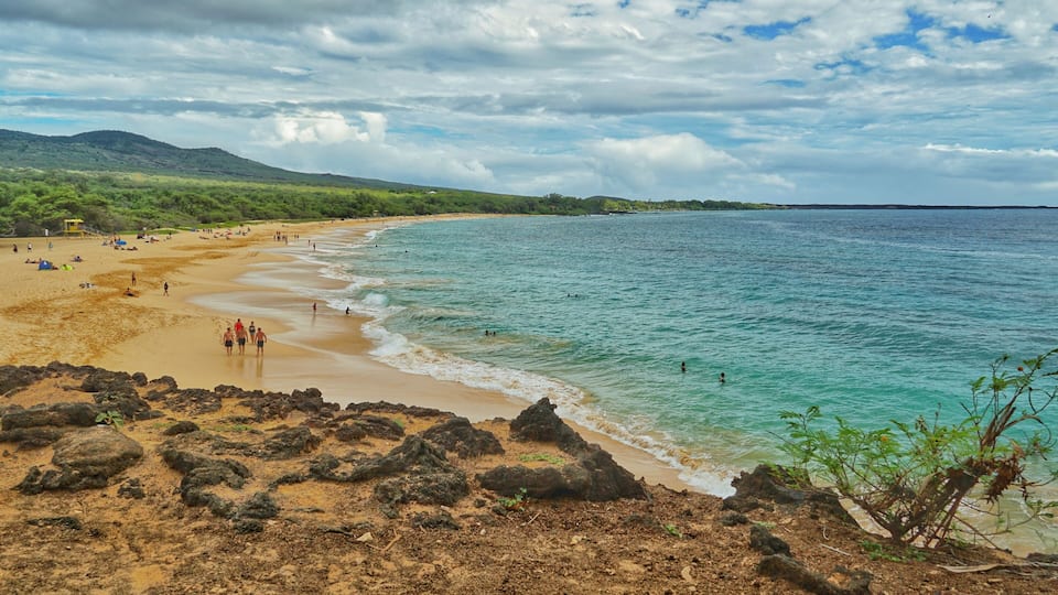 A beach with clear turquoise water, rocky shoreline, and distant mountains.