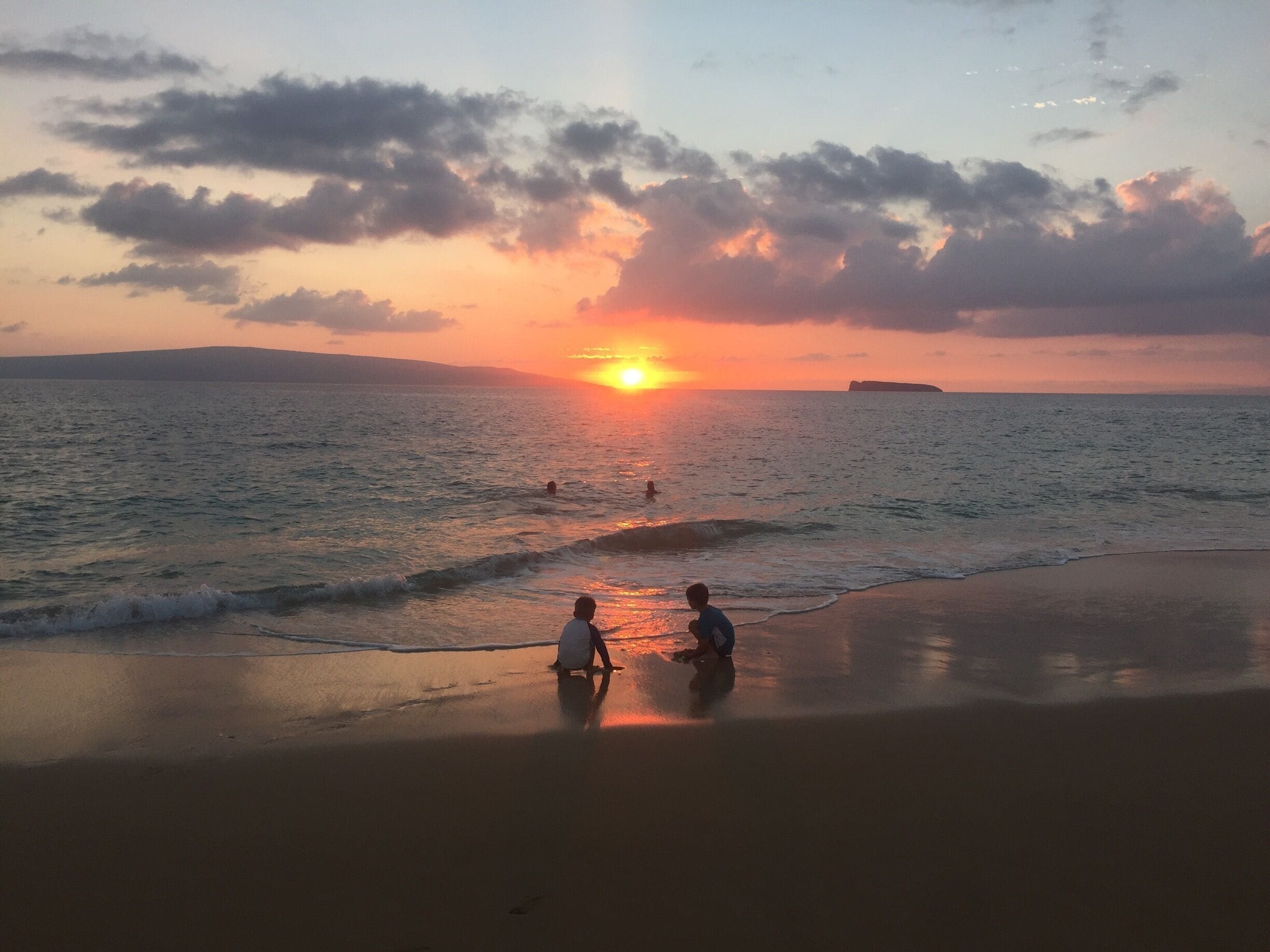 Um pôr do sol sobre o oceano, com uma praia ao primeiro plano e uma massa de terra distante.