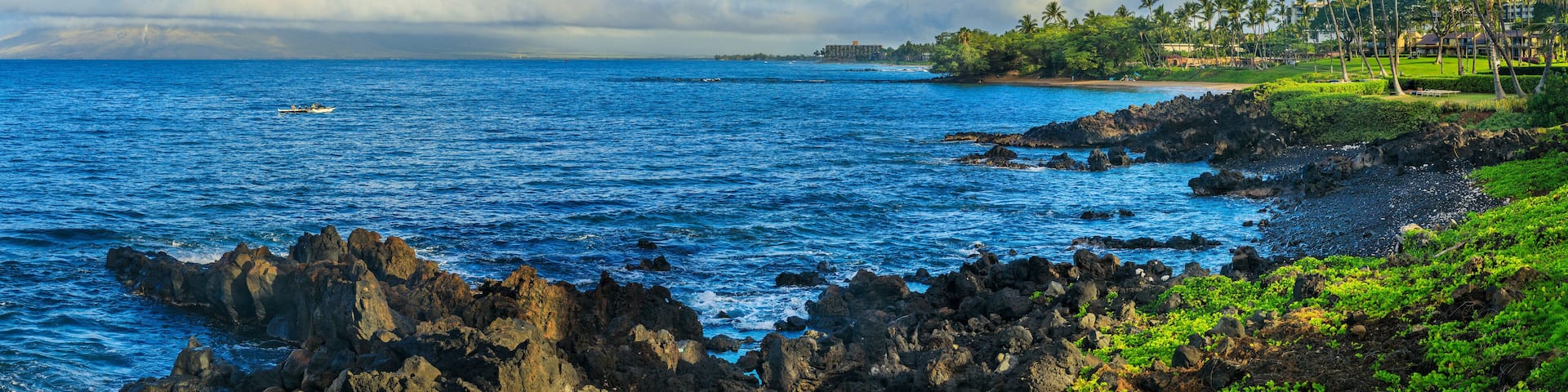 A coastal landscape with rocky shores, clear blue waters, and a distant view of a tropical resort with palm trees.