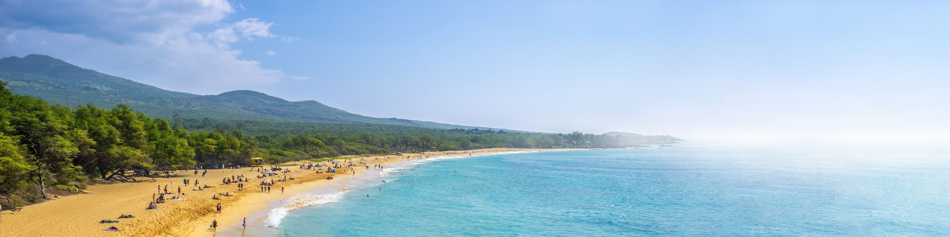 A beach with clear turquoise waters, sandy shores, and lush green hills in the background.