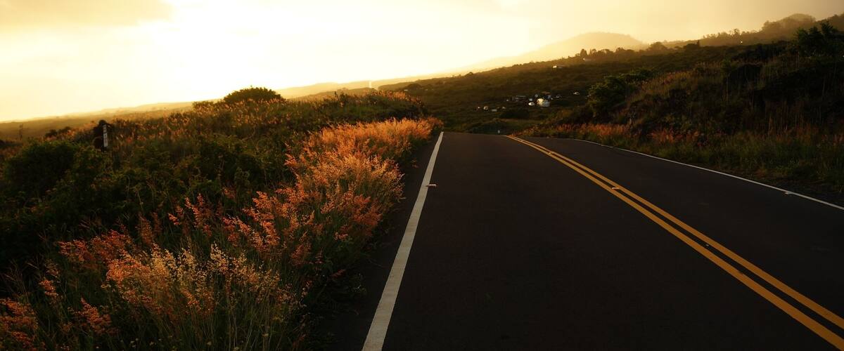 A two-lane road with a white dividing line, surrounded by vegetation and hills in the distance.