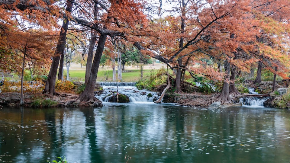 A calm river with a small waterfall surrounded by trees with autumn foliage.