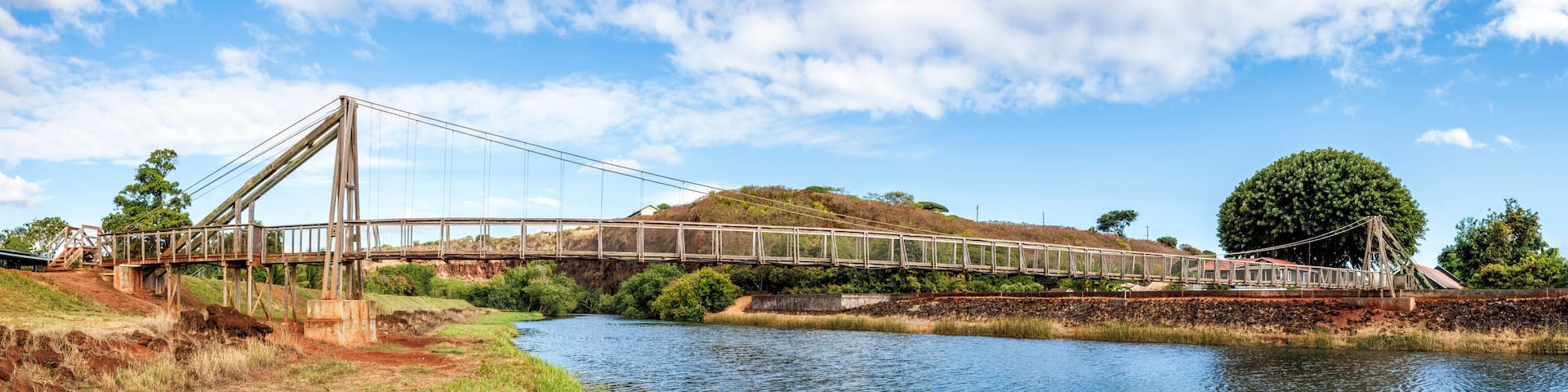 A suspension bridge over a river with a hilly landscape and trees in the background.