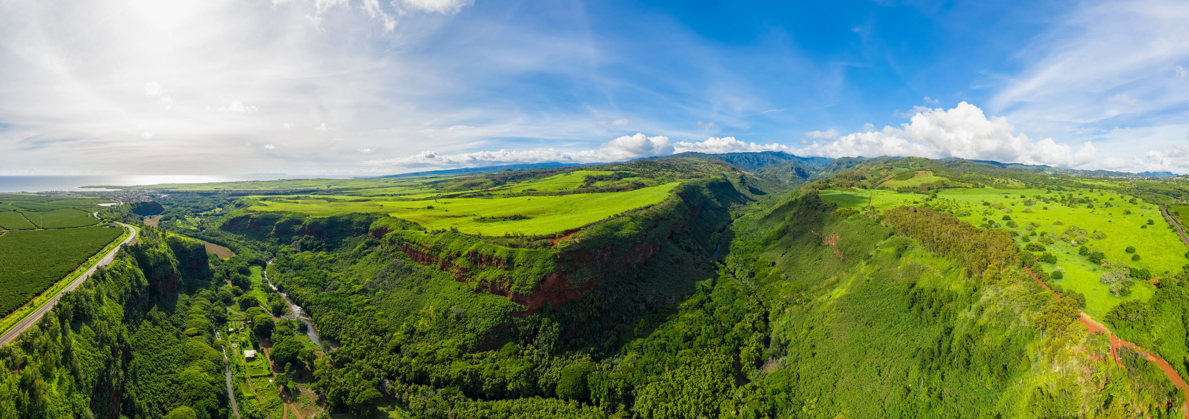 A lush green valley with a winding river, a road, and distant mountains under a partly cloudy sky.