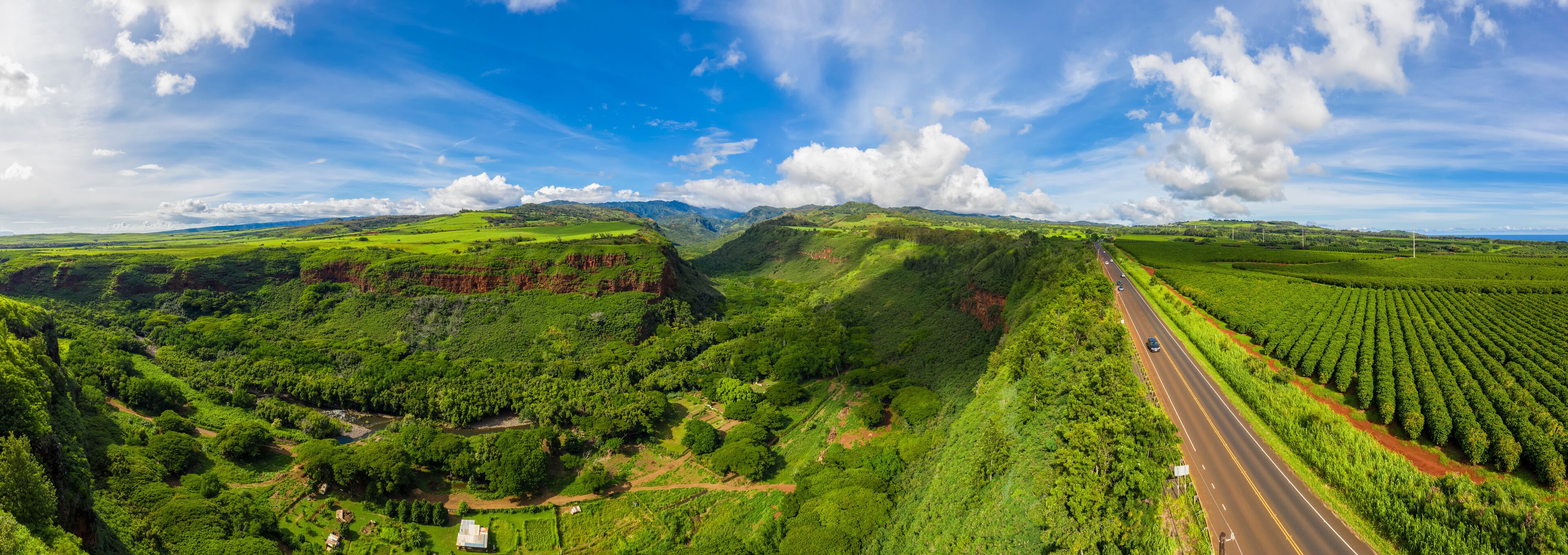 Un valle verde y exuberante con un camino sinuoso, campos y montañas distantes bajo un cielo parcialmente nublado.