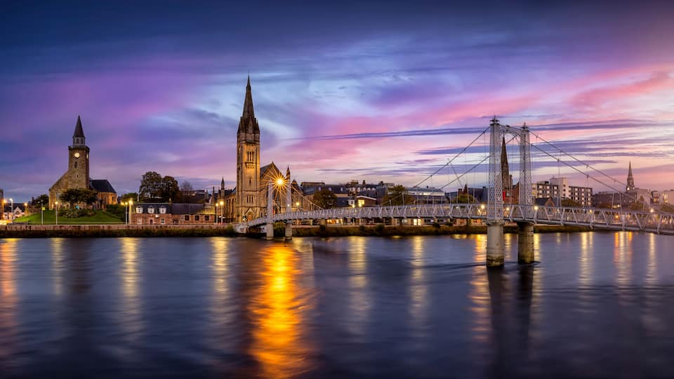 A cityscape with a historic church, a prominent clock tower, and a modern suspension bridge over a river.