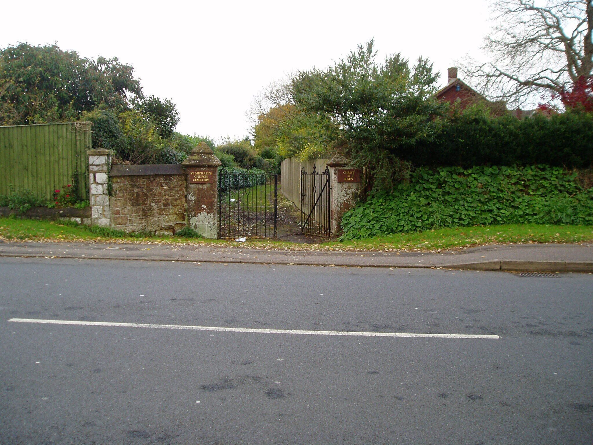 A road with a metal gate and stone walls, surrounded by greenery and a brick building.