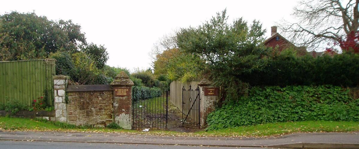 A road with a metal gate and stone walls, surrounded by greenery and a brick building.