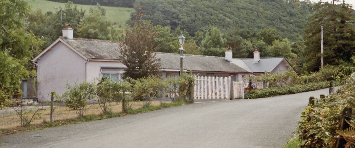 A rural house with a white picket fence, surrounded by greenery and hills.