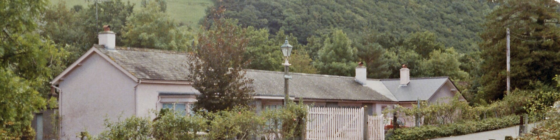A rural house with a white picket fence, surrounded by greenery and hills.