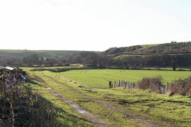A rural landscape with a dirt path, green fields, and rolling hills in the background.