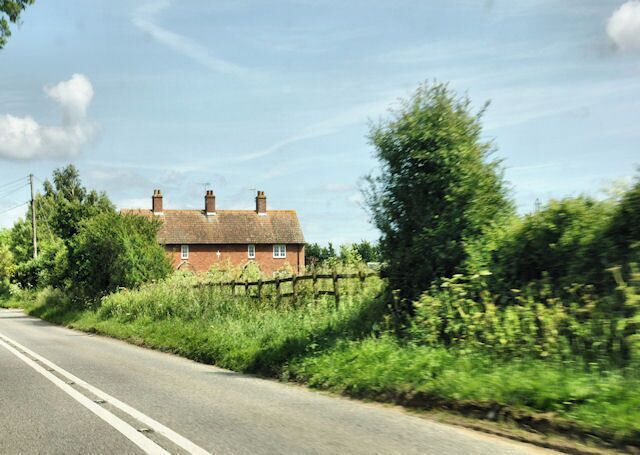A rural road with a brick house on the left, surrounded by greenery and a wooden fence.