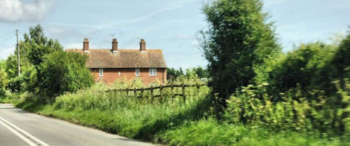 A rural road with a brick house on the left, surrounded by greenery and a wooden fence.