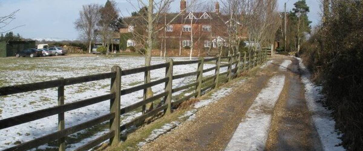 A rural road with a wooden fence, a brick house, and a snow-covered field.