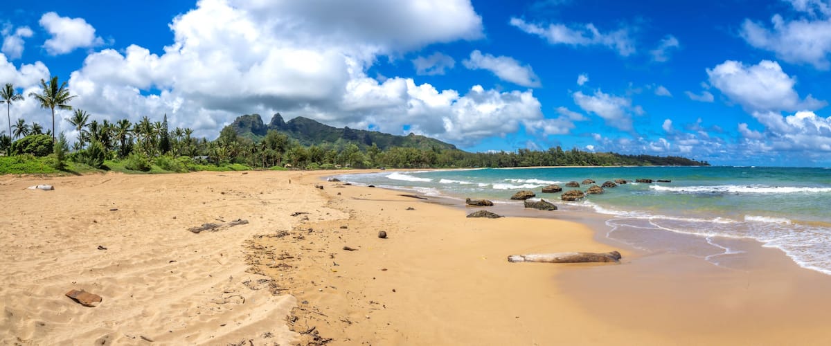 A sandy beach with clear blue water, scattered rocks, and a backdrop of lush green hills under a partly cloudy sky.
