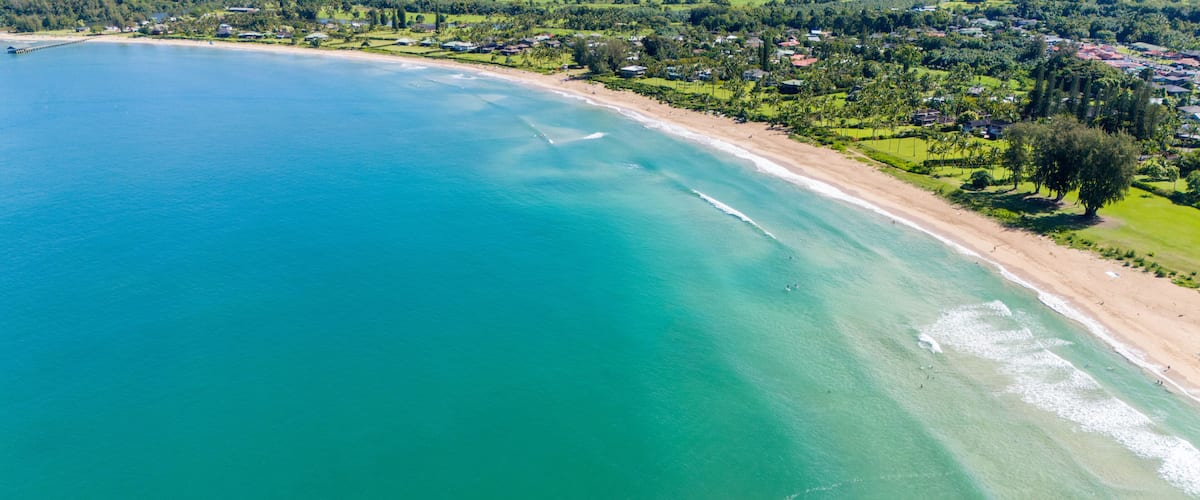 A coastal landscape with a clear blue sea, a sandy beach, and a residential area with houses and greenery.