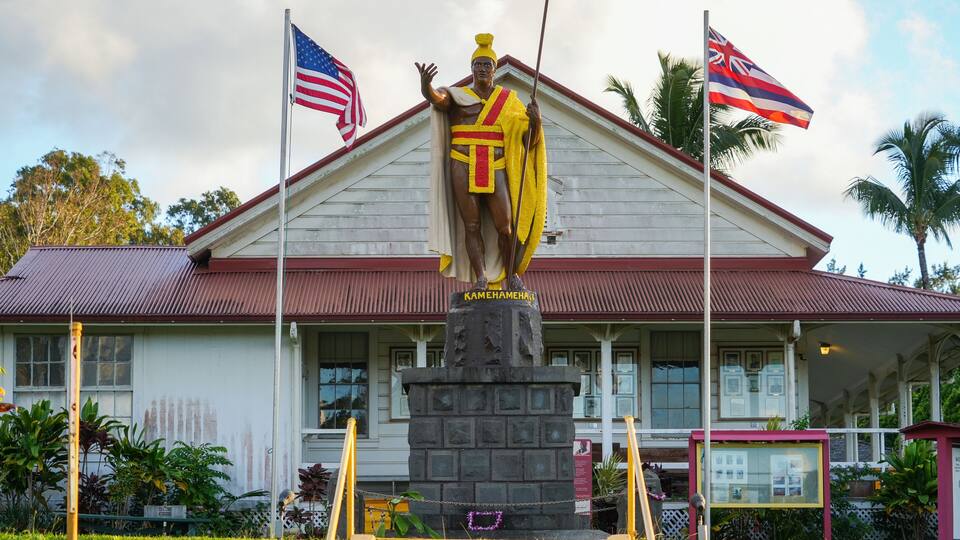 A statue of a warrior in traditional attire, with the name 'Kamehameha' inscribed on the base, in front of a building with a red roof and two flags, one of the United States and one of Hawaii.
