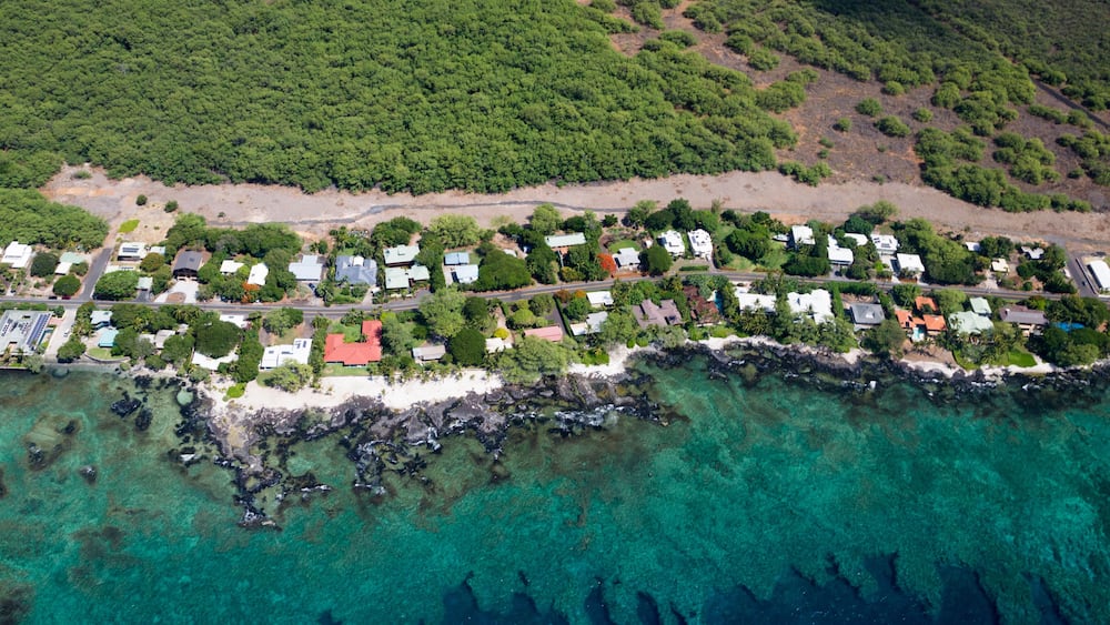 An aerial view of a coastal residential area with houses, a beach, and clear turquoise waters.