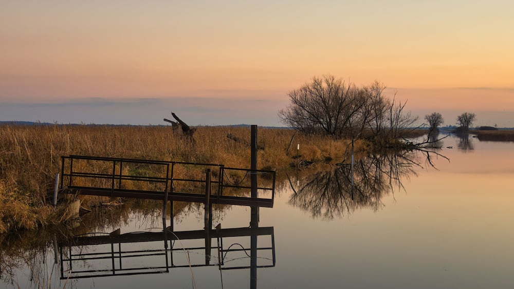 Beautiful late-Autumn landscape captured at Horicon Marsh in Dodge County, Wisconsin, as the sun sets on a peaceful river reflecting an iron pier and distant trees beneath an orange sky.