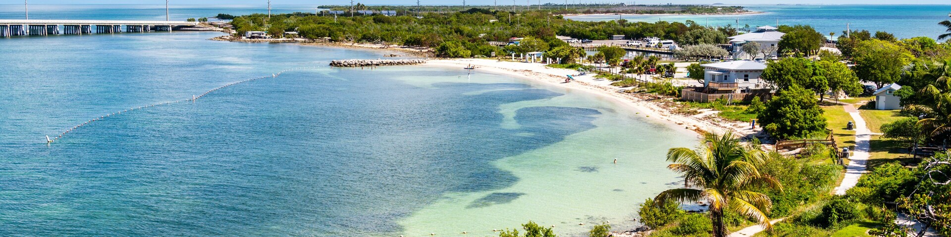 Uma vila costeira com praia, águas cristalinas, um cais e vegetação exuberante.