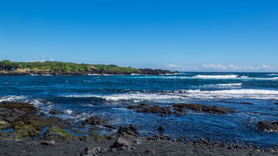 A rocky shoreline with waves crashing onto the beach and a distant view of a green, hilly landscape under a clear blue sky.