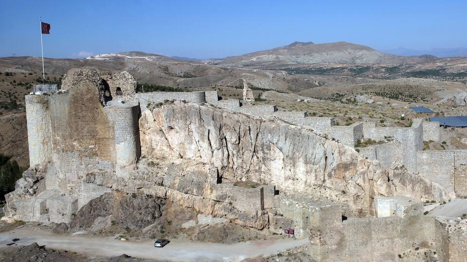An ancient stone fortress with a flag on top, surrounded by rugged hills and a clear sky.