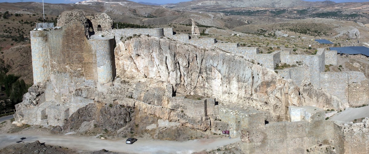 An ancient stone fortress with a flag on top, surrounded by rugged hills and a clear sky.