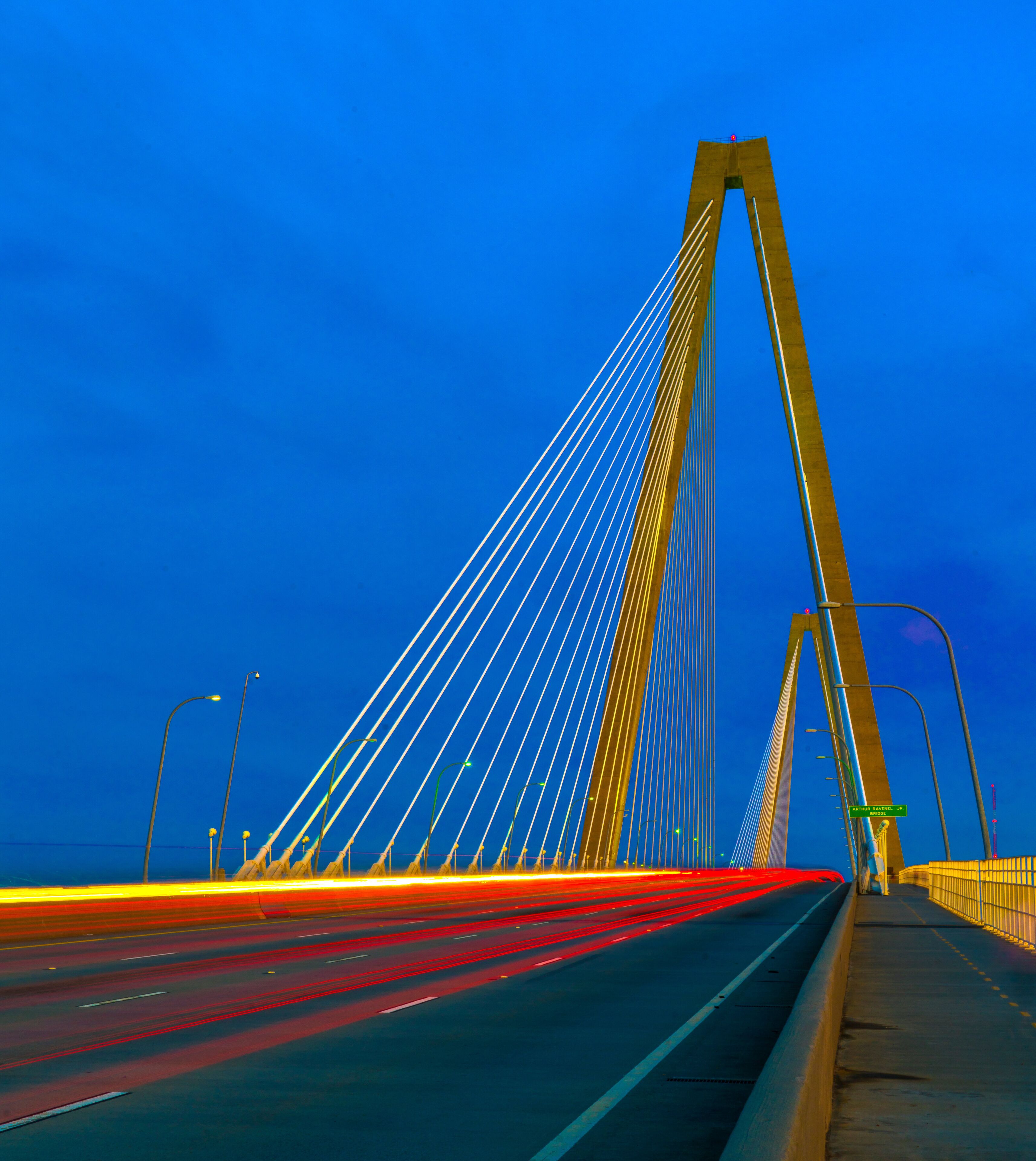 A bridge with a tall central tower and cables, a road with moving lights, and a clear blue sky.