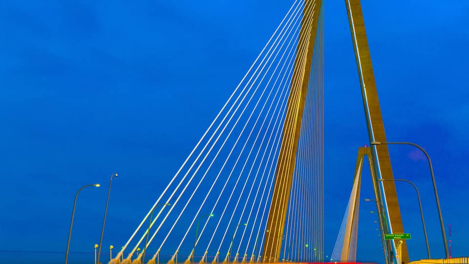 A bridge with a tall central tower and cables, a road with moving lights, and a clear blue sky.