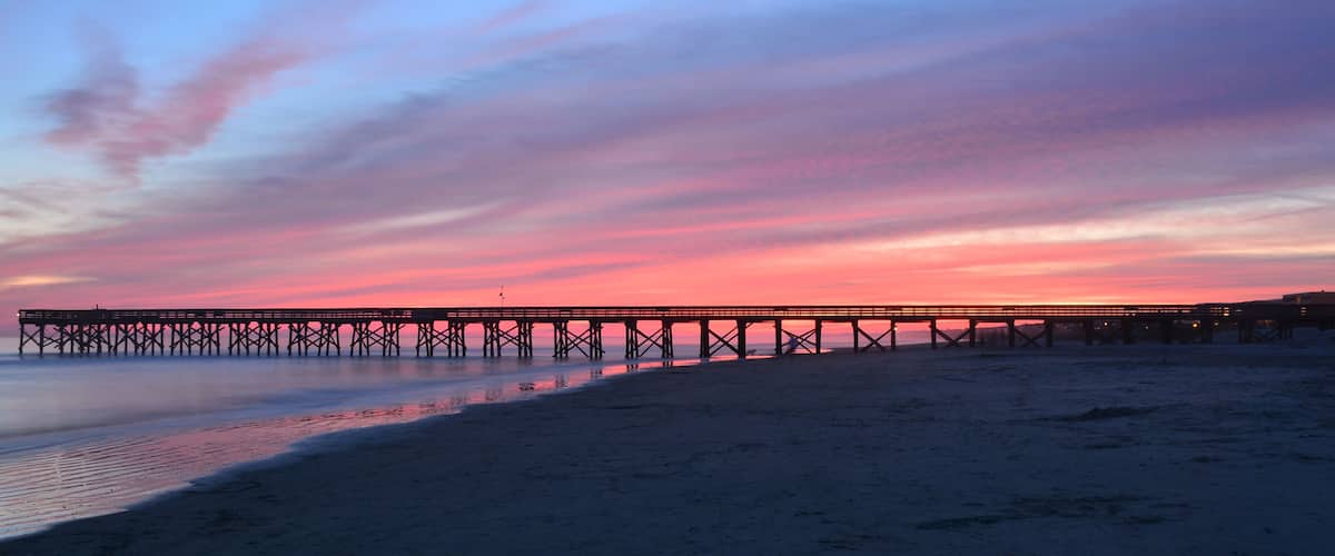 A pier extending into calm waters at sunset with a pink and blue sky.