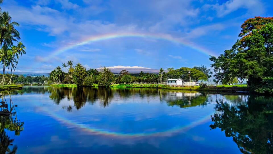 A lake with a rainbow, palm trees, a white building, and a mountain in the background.
