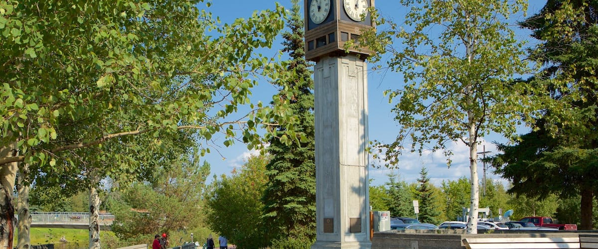 A tall clock tower with Roman numerals, surrounded by trees and a bridge.