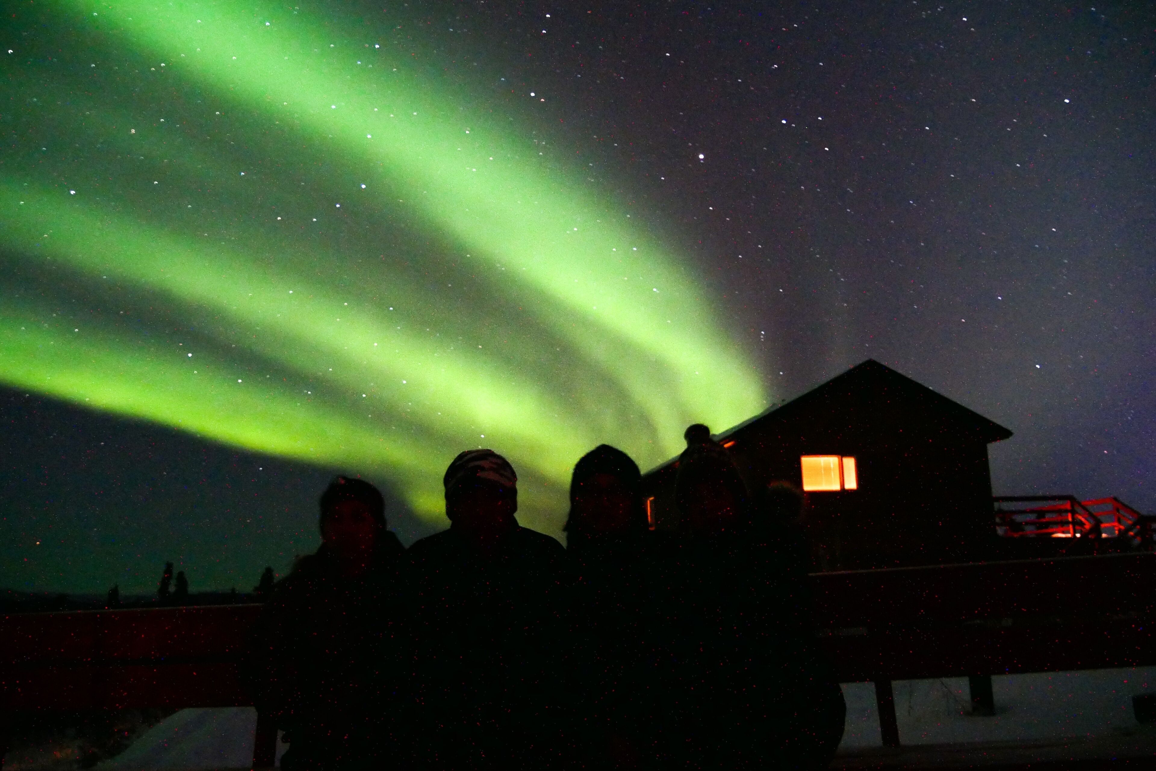 The Northern Lights over a snowy landscape with a cabin and people observing.