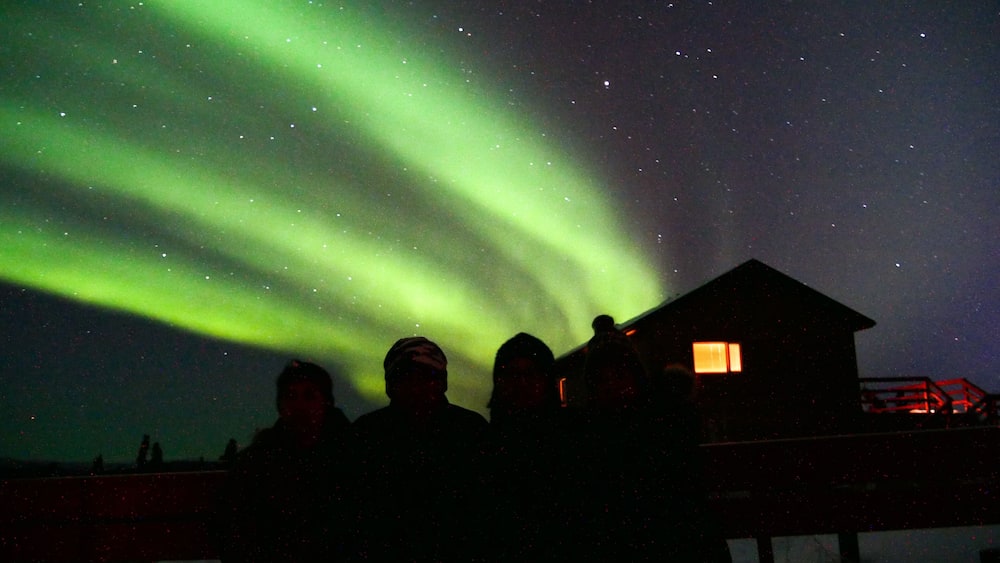 The Northern Lights over a snowy landscape with a cabin and people observing.
