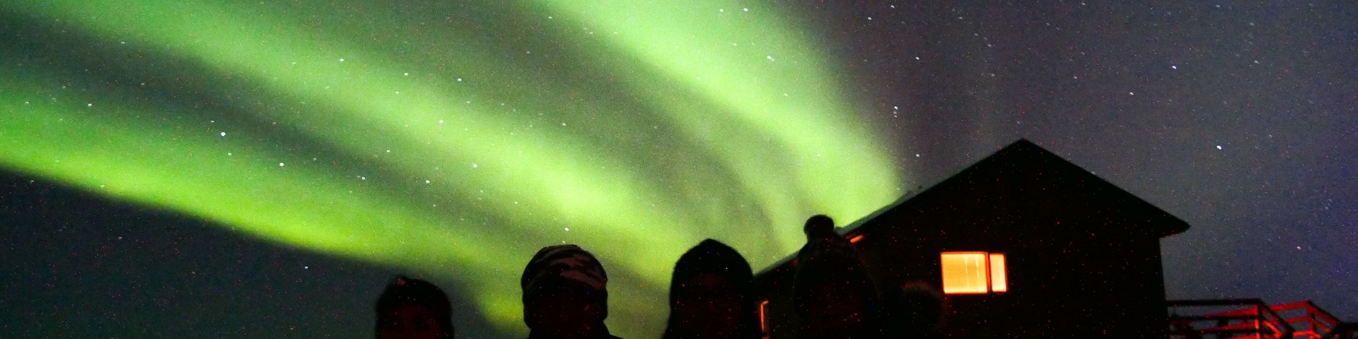 Auroras boreales sobre un paisaje nevado con una cabaña y gente observando.
