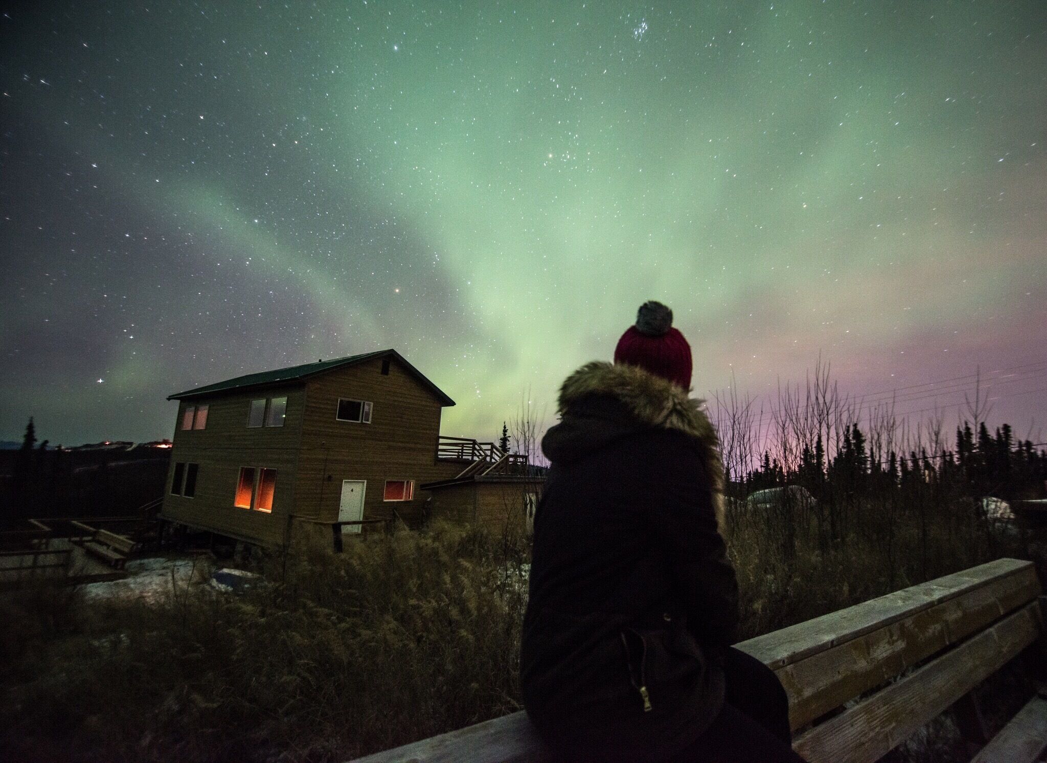 A person in a red jacket and fur-lined hood overlooking a wooden house with a lit window, under a starry night sky with the aurora borealis.