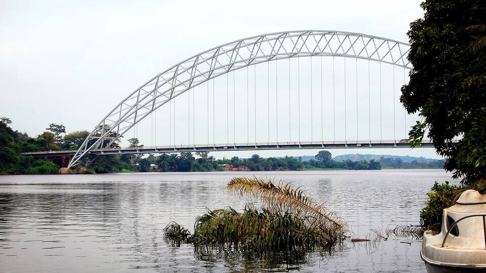 A large steel bridge spanning a river with dense greenery and trees in the background.