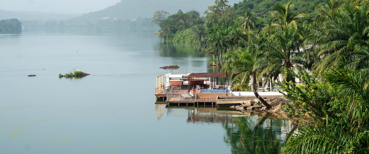 A calm lake with a wooden dock and a small structure, surrounded by lush greenery and palm trees.