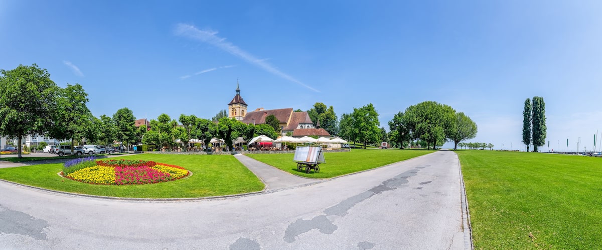 A park with a flower bed, a church, and sailboats in the background.