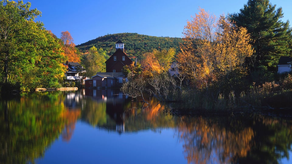 A serene lake with autumn foliage, a red building, and a hill in the background.