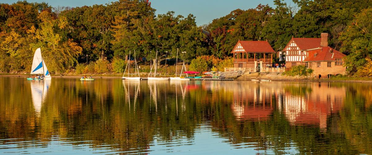 Jamaica Pond view across the water of the boat house and music building. Boats are in the water. A reflection of the buildings are on the water. There is a blue sky. Taken during the golden hour.