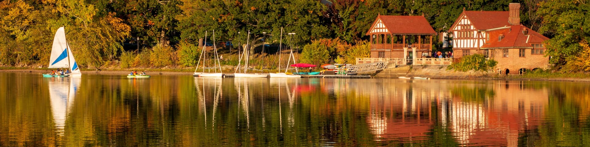 A calm lake with a sailboat, a traditional house, and dense trees in the background.