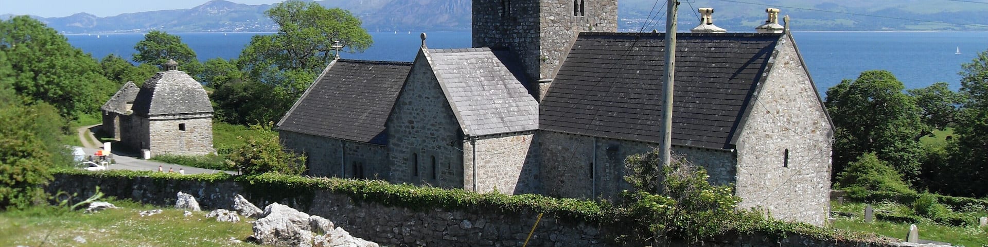 A stone church with a bell tower and a smaller building, surrounded by greenery and overlooking a body of water.