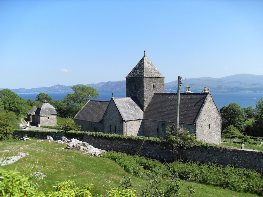 A stone church with a bell tower and a smaller building, surrounded by greenery and overlooking a body of water.