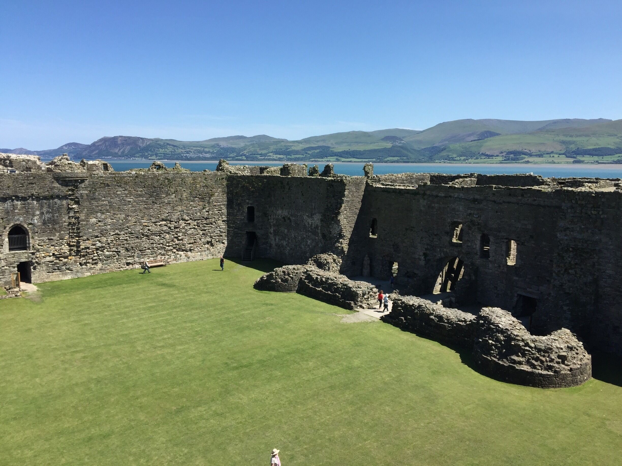 A historic stone castle with a grassy courtyard and a scenic view of hills and a body of water in the background.