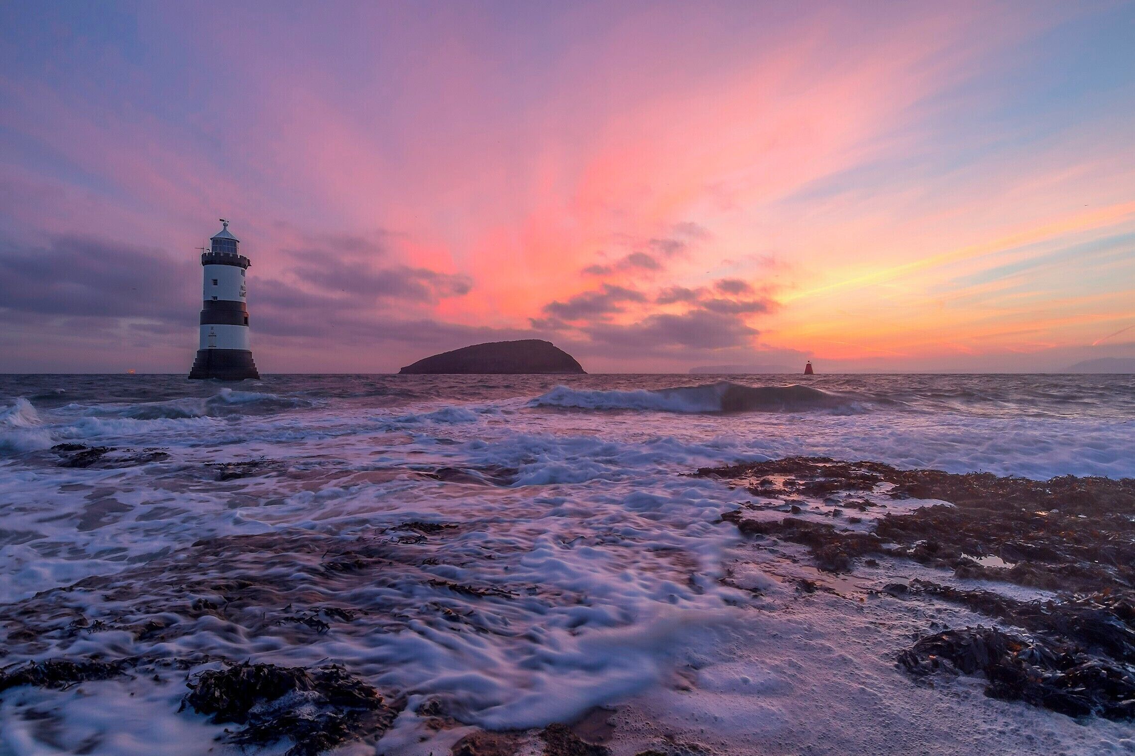 A lighthouse, rocky shoreline, and a distant island at sunset.